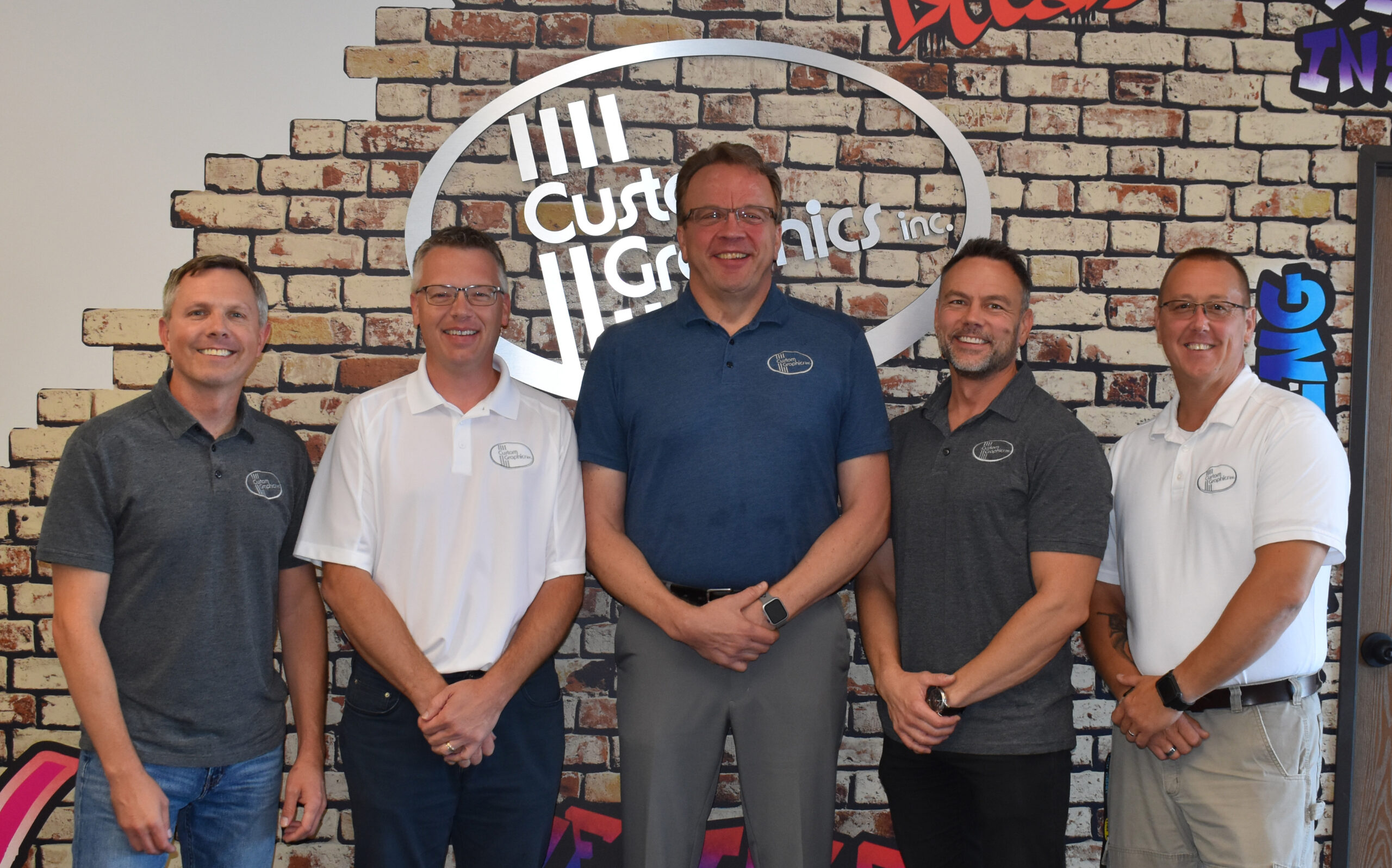The team of five salesmen at Custom Graphics stand in a line in front of a wall wrapped in a faux brick vinyl pattern with a brushed aluminum channel letter logo.